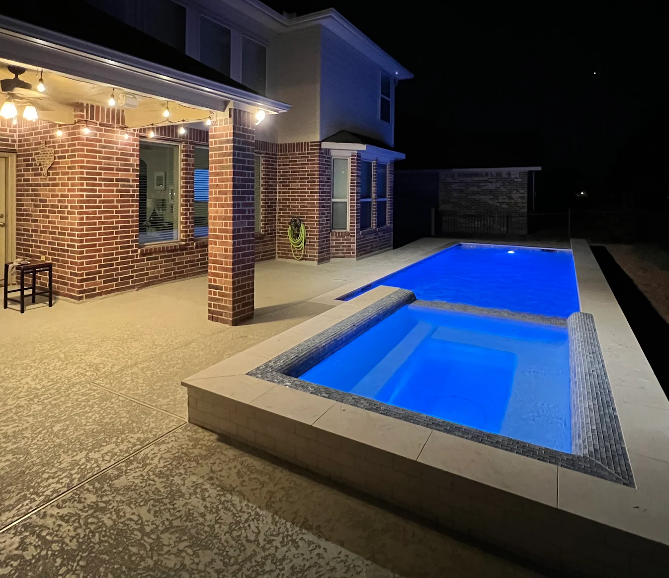 Modern backyard with illuminated blue swimming pool and cozy patio lights at night.